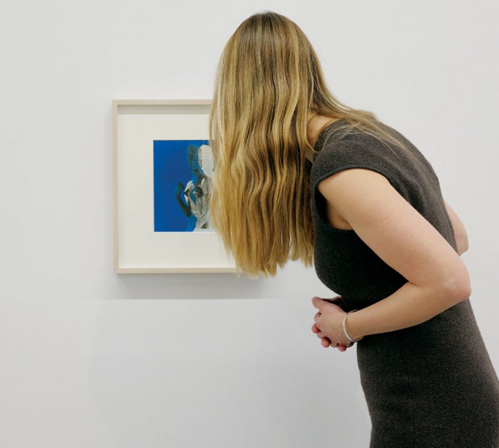 Woman has back turned to camera as she views a framed art painting on white wall.