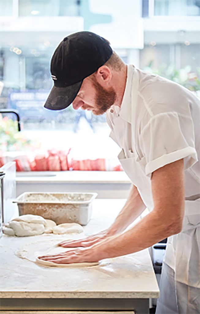 Employee flattening pizza dough by hand.